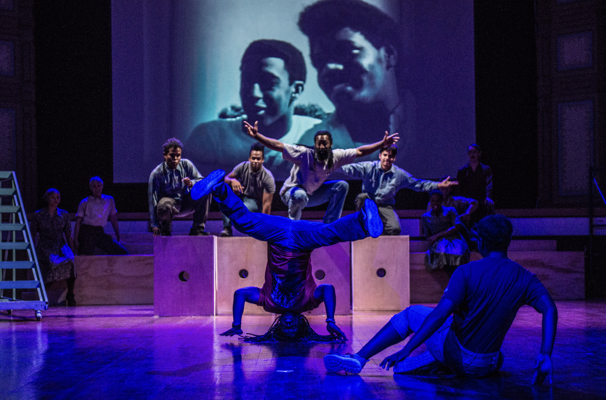 A handful of dancers perform on stage in front of a projected image of two African-American people on a wall. 